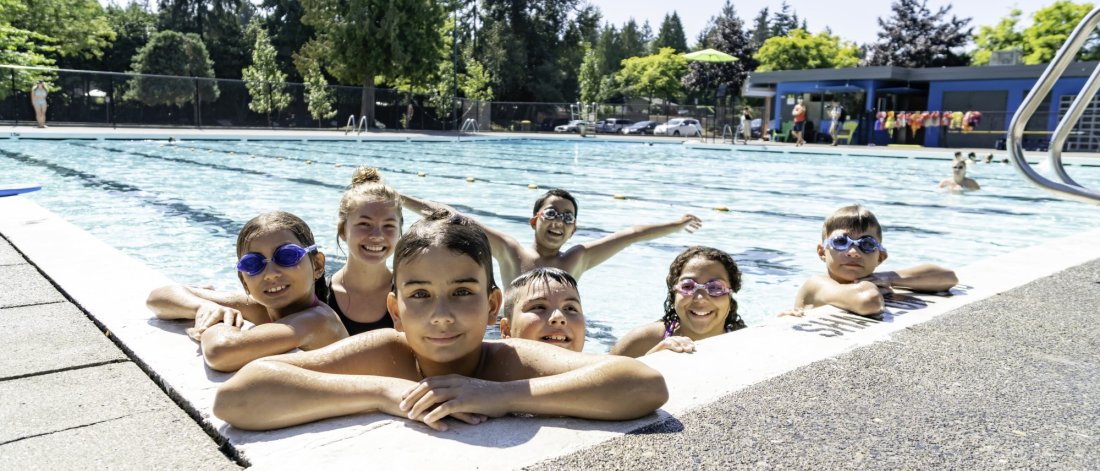 Group of children with swim goggles, smiling and enjoying themselves at an outdoor pool on a sunny day.