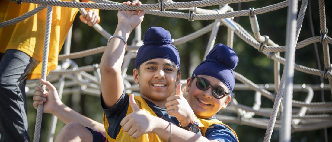  Two young boys in turbans giving thumbs up while climbing on a rope structure at a playground.