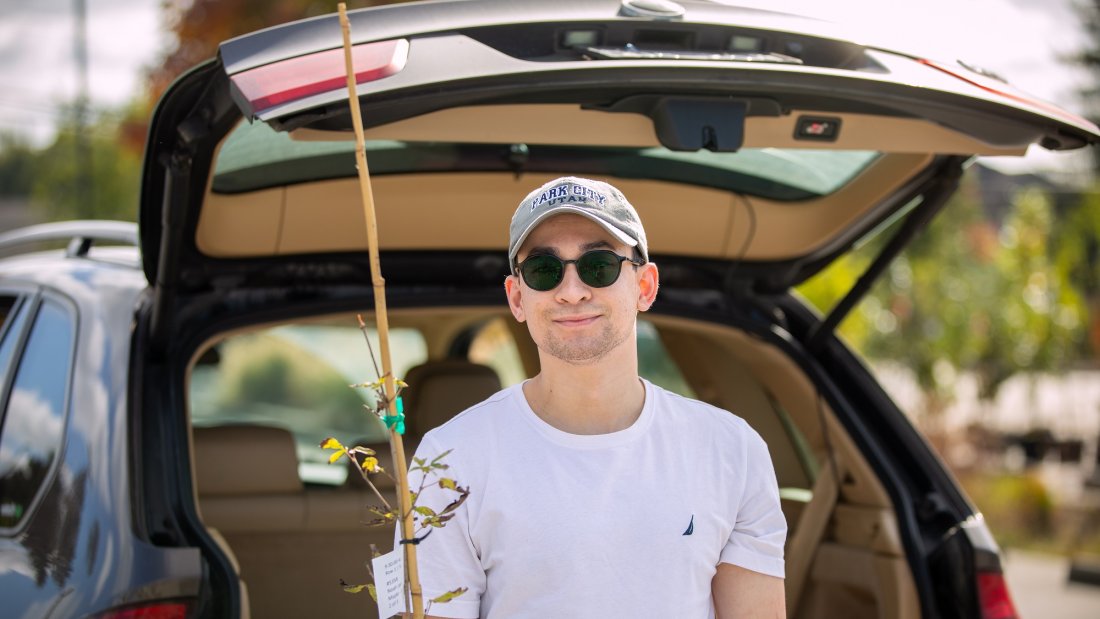 Person smiling holding small tree, next to vehicle with trunk open