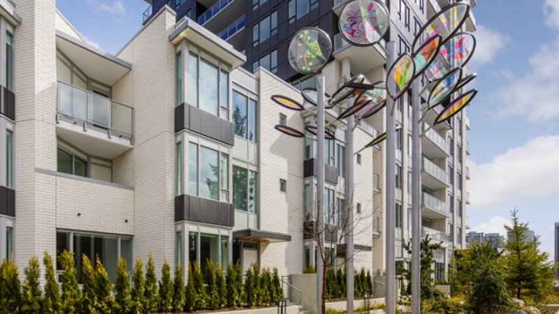 A vibrant kinetic sculpture with colorful, translucent elements stands in front of a modern apartment building