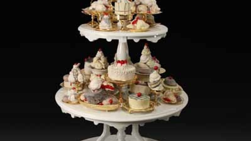 A three-tiered cake stand made of white ceramics are covered with pastries that could be found in an afternoon tea. The sweet treats are small and placed on top of mini trays and holding dishes with a golden bronze sheen. Whipped cream dotted by red berries can be seen. 