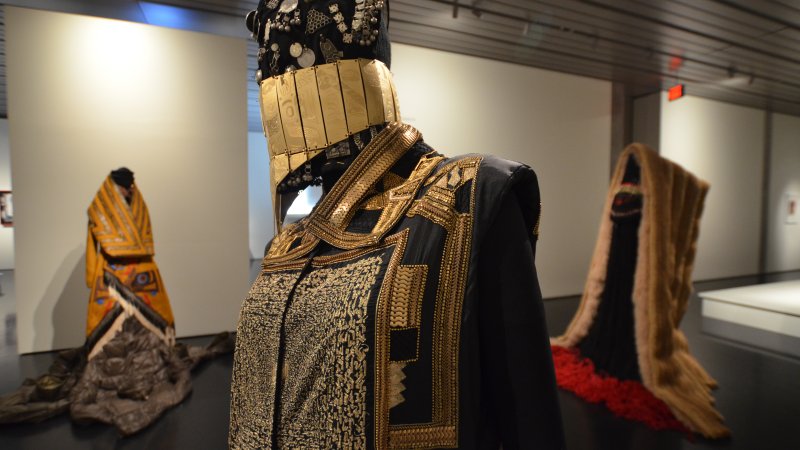 A mannequin can be seen in profile from the torso up. It wears a draped garment that is black with brass and gold decorations. There a square design covering the chest. The lapels are in brass gold scales. There is a golden mouth covering and a black hat with silver details on the head.  