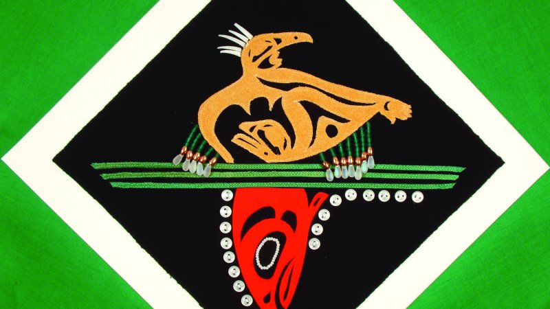 A detail of a quilt that shows a unique hexagonal block. The background is forest green. There is a white diamond shape and inside this shape is a black diamond shape. There is a yellow bird and three green, horizontal lines. Below the lines is a fish head. The fish is depicted with red colours. 