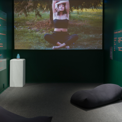 Two green walls with text and photographs frame a space with two yogibos. A film plays on the wall in between the two greens where a woman sitting cross-legged on grass in nature is shown. She has her elbows bent overhead, one hand cups her jaw, the other hand is behind her back.