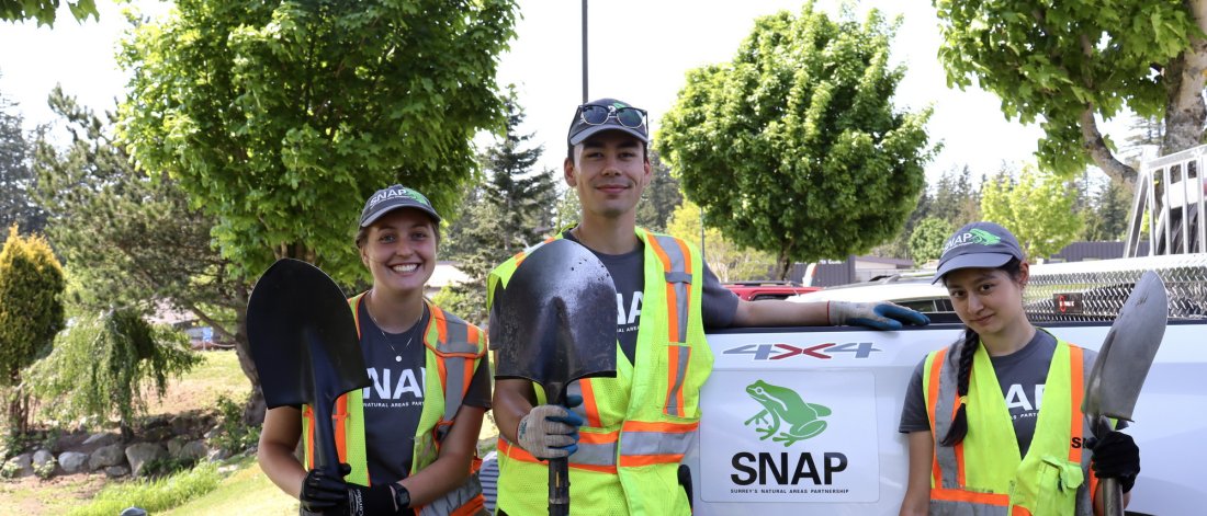 three people holding shovels, wearing high-visibility vests with park in background