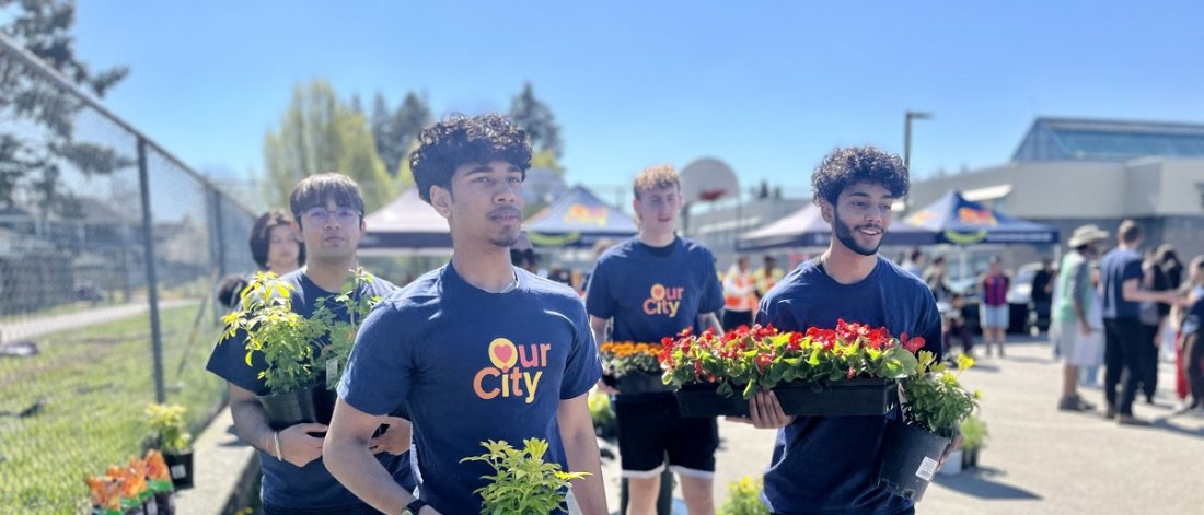 Group of volunteers wearing "Our City" t-shirts carrying plants at an outdoor community event.
