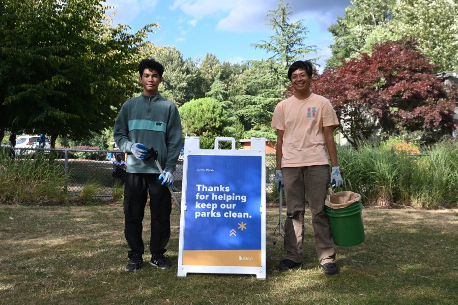 two youth with litter pickers and a bucket stand next to a park cleanup sign