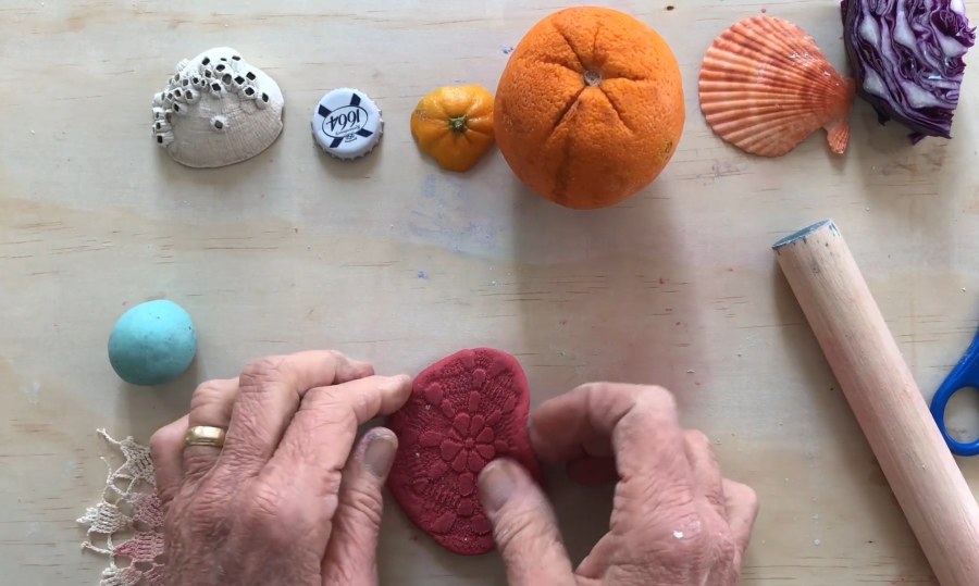 Flatlay of a person's hands holding onto a sphere of red dough with a flower emblem. There is a turquoise blue ball, a seashell, a bottle cap, orange rind, an orange, an orange shell, and a piece of purple cabbage in front of the hands.