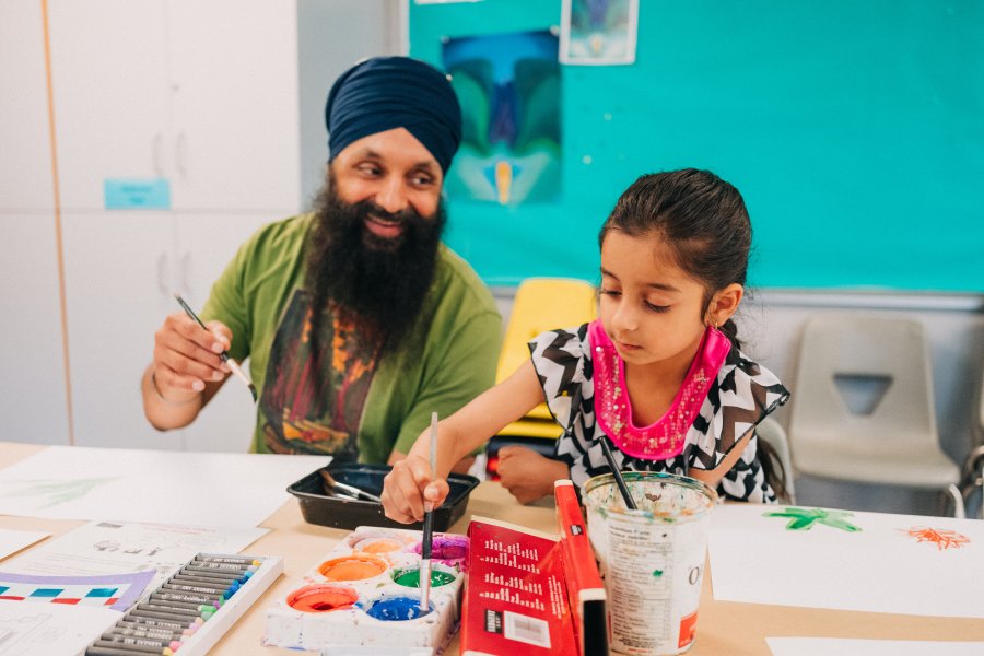 A man with a turban holds a paintbrush, he looks with love at a child who is also holding a paintbrush. She is dipping it in blue paint. They are seated in an art classroom.