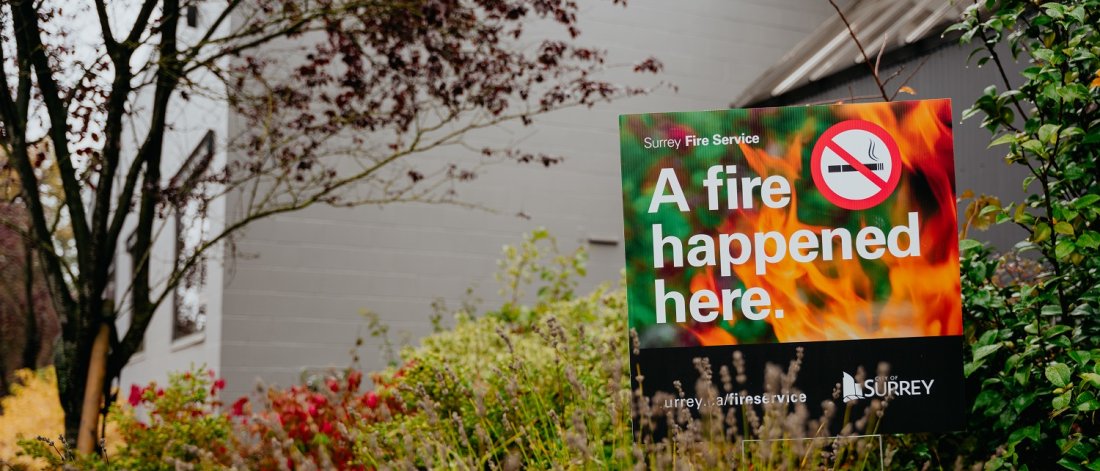A sign by Surrey Fire Service with a warning message "A fire happened here." against a backdrop of trees and bushes.