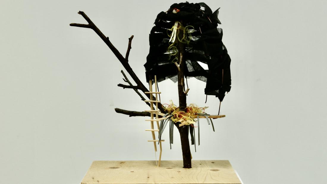 Sculpture of a tree branch from which a miniature stick ladder and various textiles are hung.