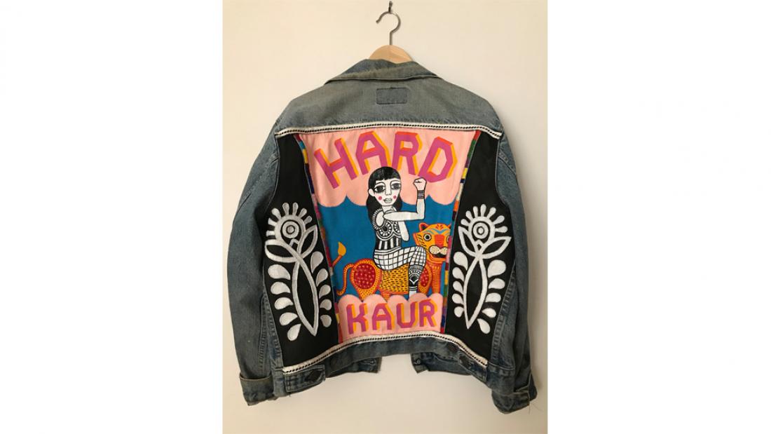 Photograph of the back of a denim jacket with the words "Hard Kaur" on it and a South Asian woman painted in the middle flexing her arm.