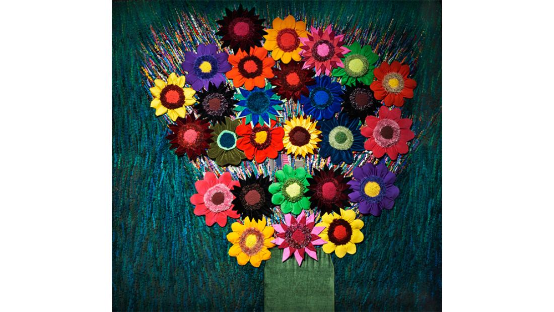 A tapestry of brightly coloured flowers using recycled materials arranged in a vase against a dark green background.