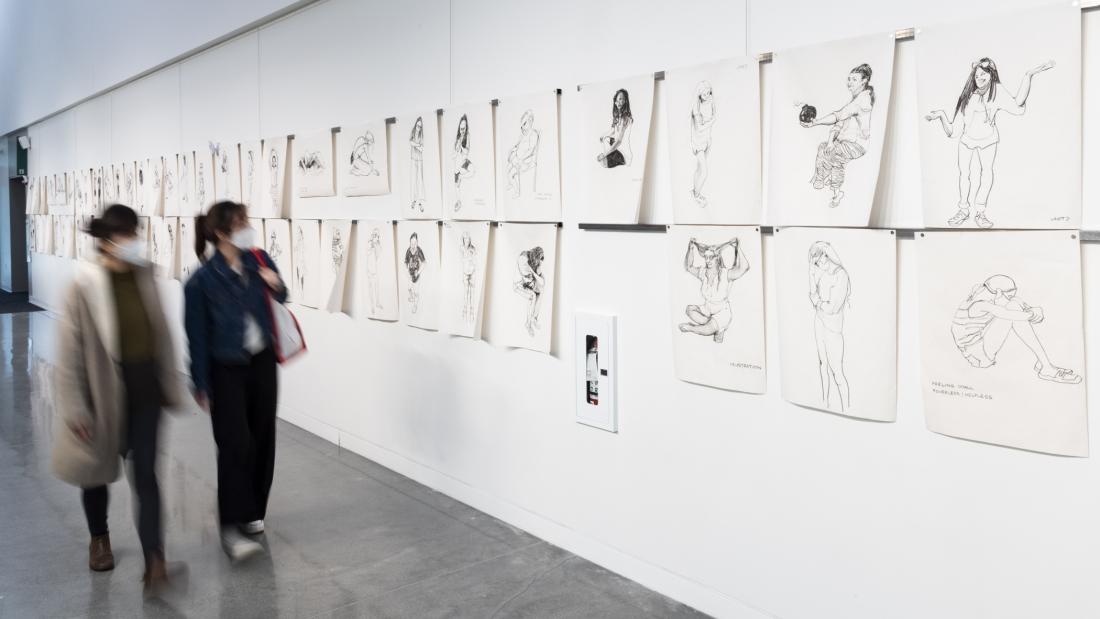 A long gallery wall with life-drawing images hung on it and two people walking towards the camera, looking at the drawings.