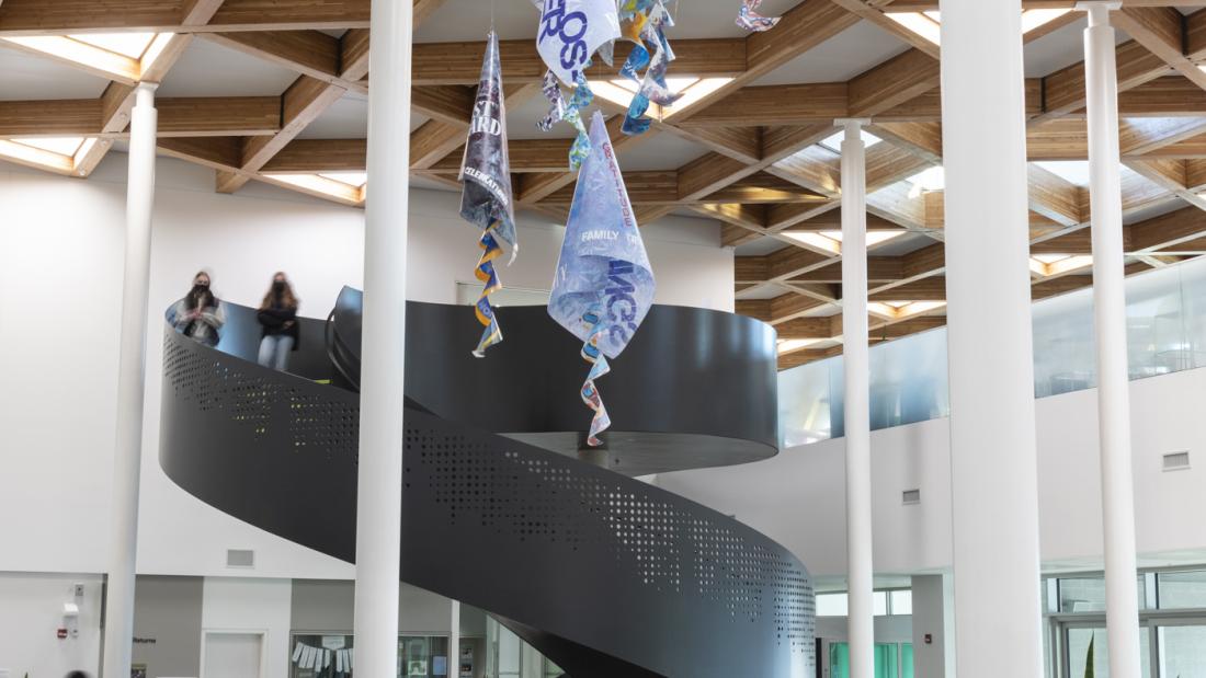 Image of a hanging sculpture made from acetate sheets rolled into cones and hung in spirals, painted in shades of blue with words stenciled on. This image shows the sculpture from far away, with a large spiral staircase in the background.