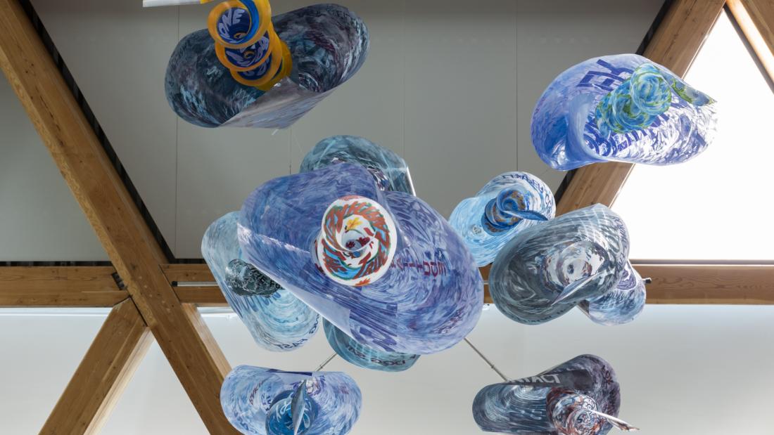 Image of a hanging sculpture made from acetate sheets rolled into cones and hung in spirals, painted in shades of blue with words stenciled on. Photo from underneath the sculpture looking straight up at it.