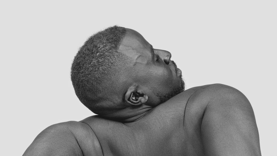 A black and white photograph of the back of a Black man without a shirt, leaning back on his arms and head tilted to the right.