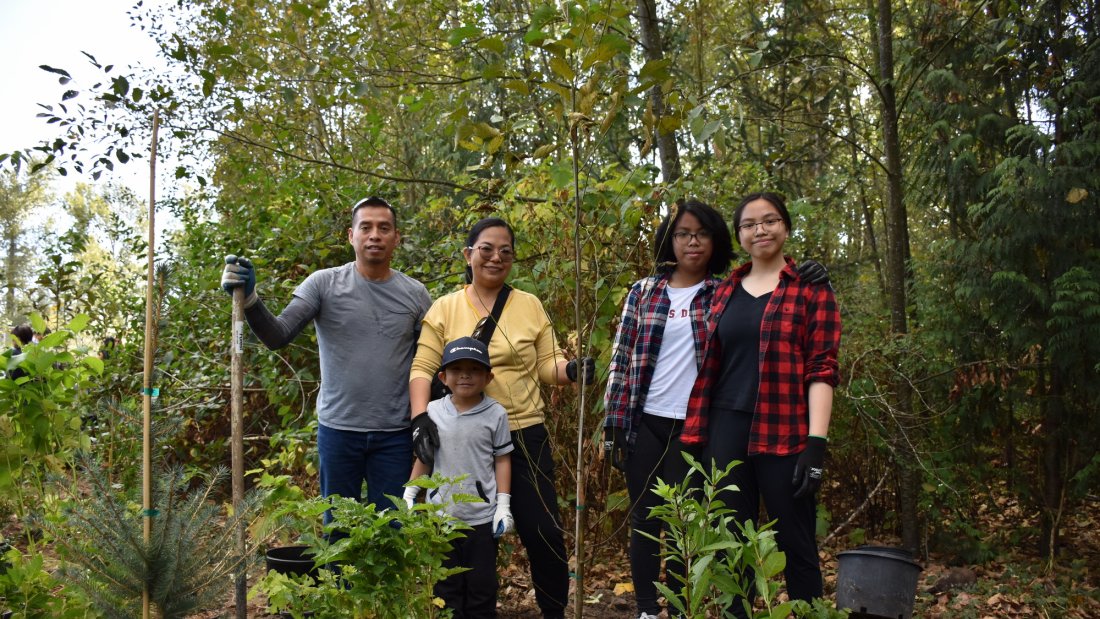 family of five holding shovel and standing next to newly planted tree
