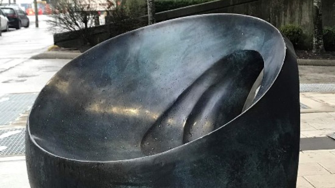  A dark, polished, seed-shaped sculpture with a spiraling groove sits on a wet sidewalk, with bare trees and a street in the background.