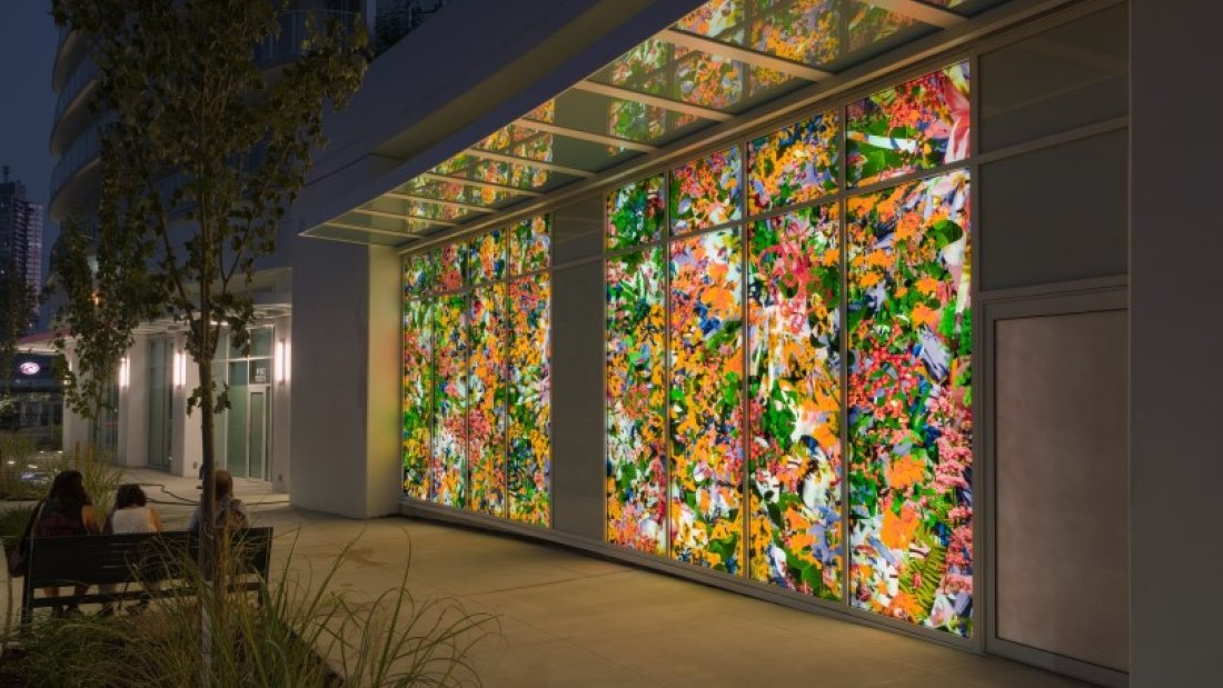  A brightly lit floral art installation on an urban building window at night, with people seated nearby.