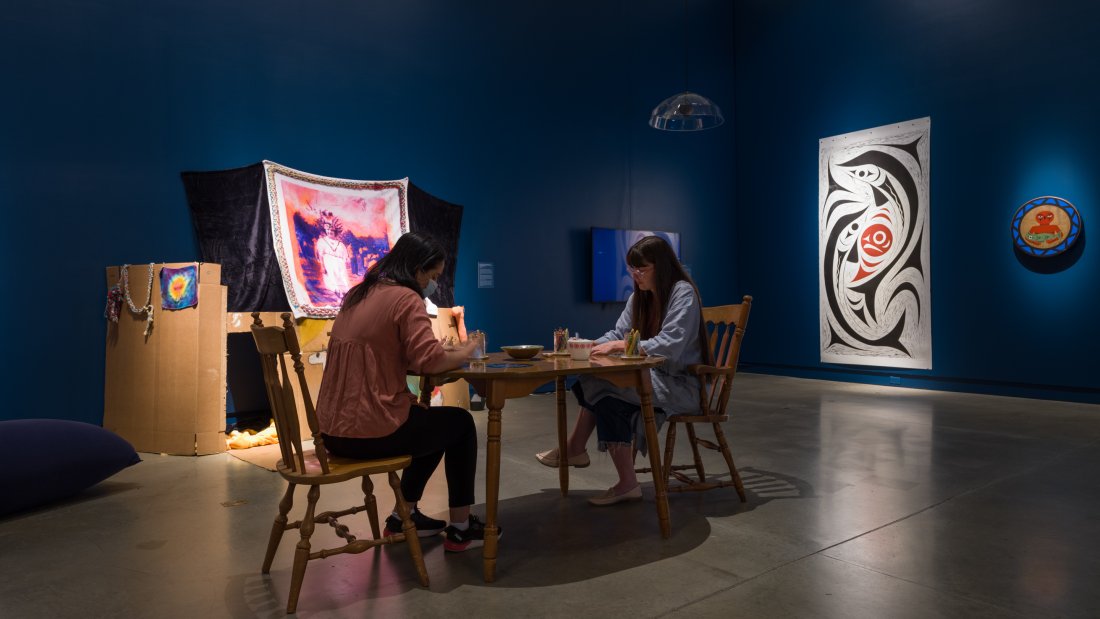 Two people sit at a kitchen table drawing in the middle of an art exhibition.