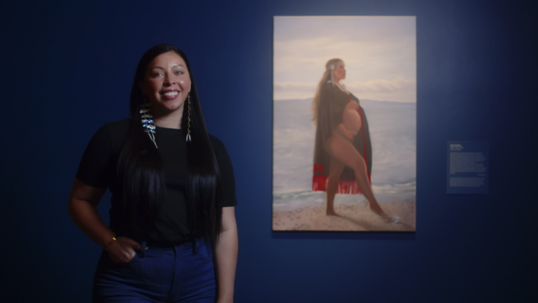 A person stands next to her painting of a woman in regalia.