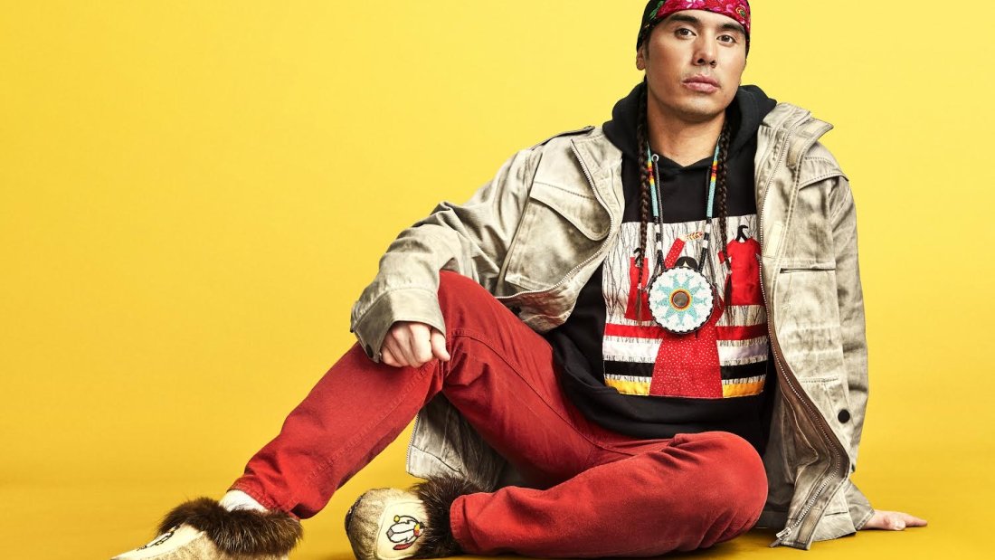 A man dressed in a blend of modern and traditional Indigenous clothing sits against a yellow background.