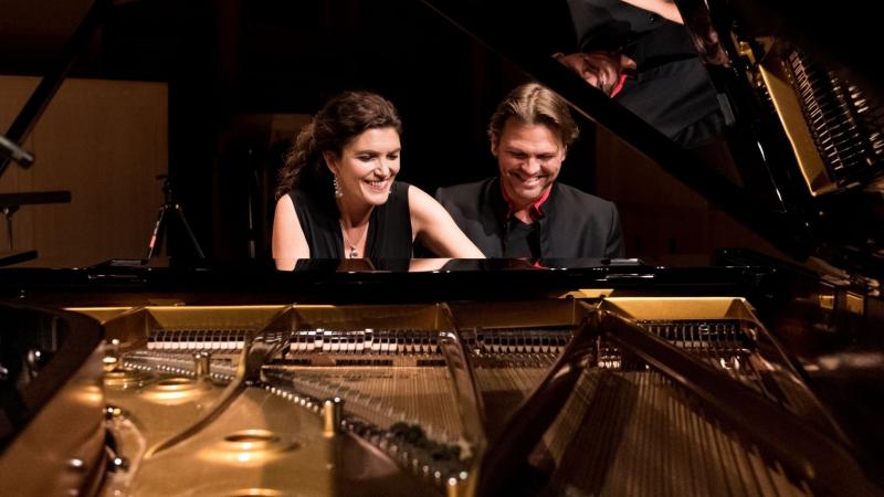 The Begmann Duo sit side by side smiling and playing a grand piano.