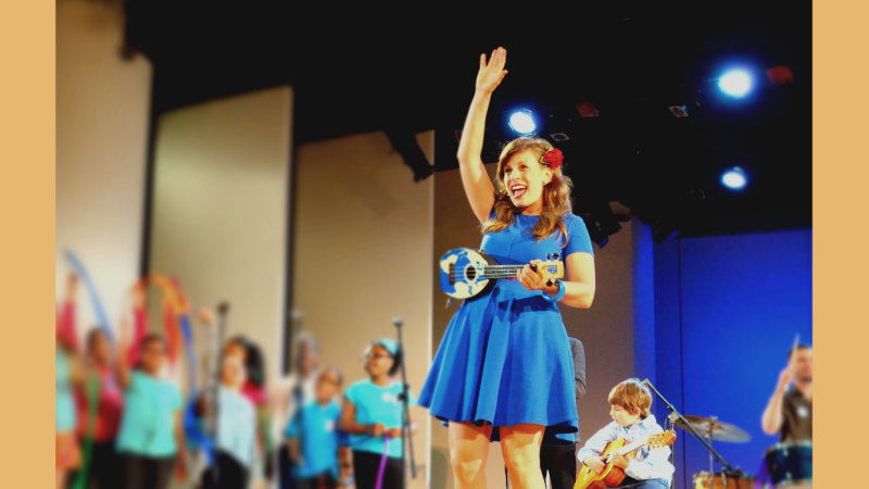 Mill wears a blue dress and holds a blue ukulele and waves at the audience
