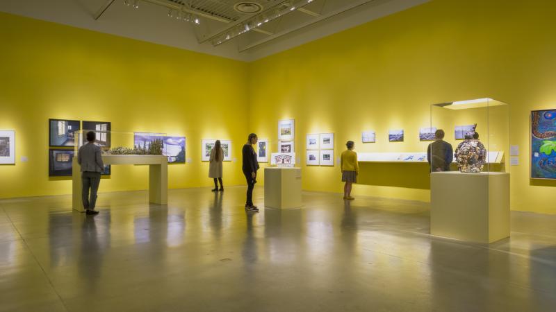 Several visitors stand at various spots around an art gallery painted with yellow walls and showing variety of art.