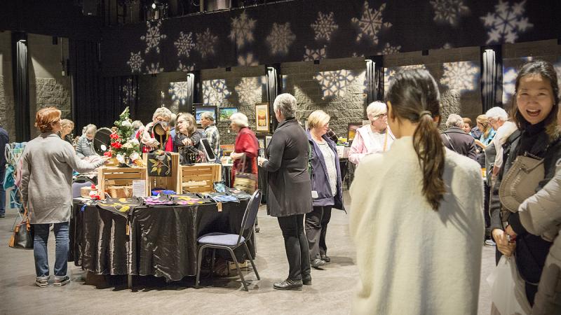 Group of shoppers standing around tables at a holiday market inside a black box theatre.