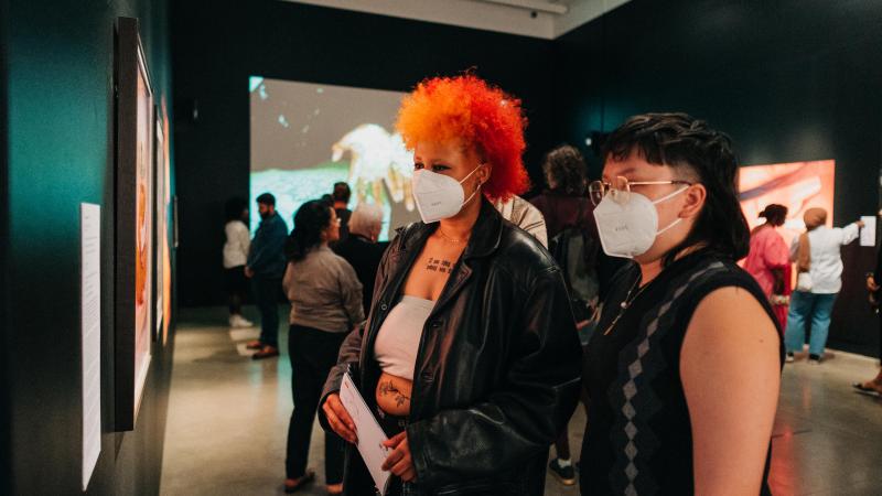 Two visitors wearing medical masks look at paintings on a wall of an art gallery with other visitors viewing art behind them.