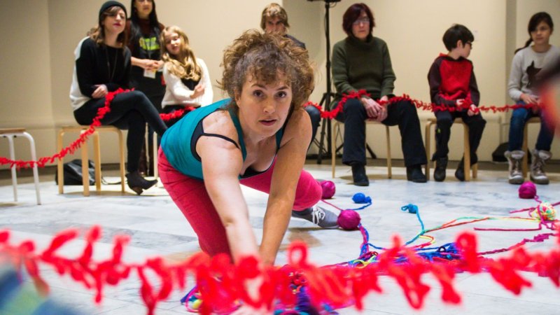 A woman is surrounded by a long stretches of red, blue and purple yarn as she looks at the camera as she dances low to the ground, the audience is sitting in a circle