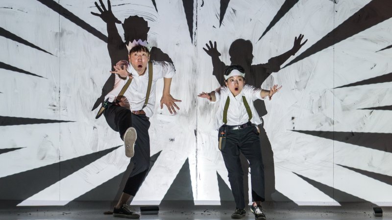 Two performers for show Doodle POP stand on stage with arms outstretched in the centre of a background projection of black and white lines.