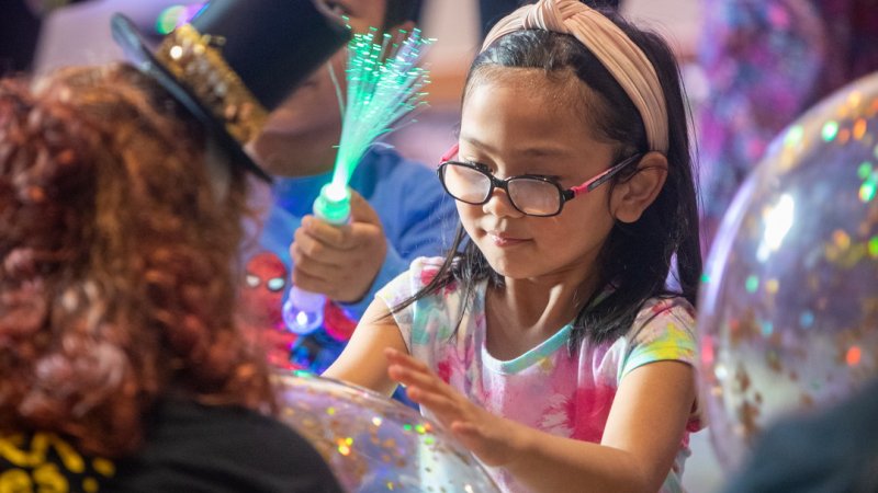 Young girl holding a glitter beach ball and boy holding a glow wand in a busy background of kids dancing 