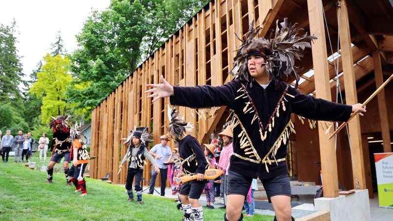 youth indigenous dancers in traditional clothing perform on grass in front of new lumber indigenous learning house