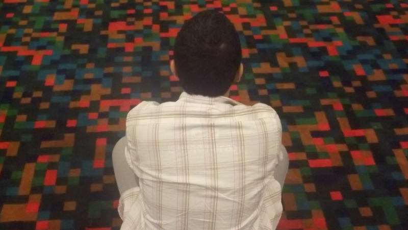 A person with short dark hair faces away from the viewer. They are wearing a white shirt and are crouched over a carpet with geometric square patterns.