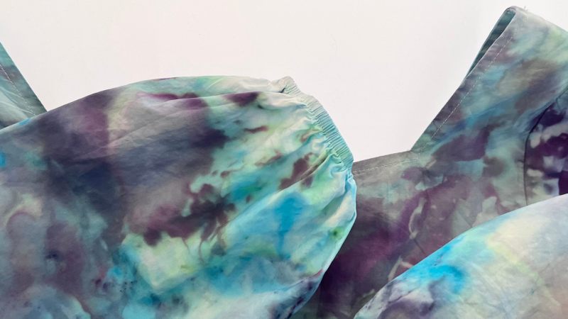 Overhead shot of a tie dye blouse of dark blue and turquoise colours. The he sleeves are folded across the chest. 