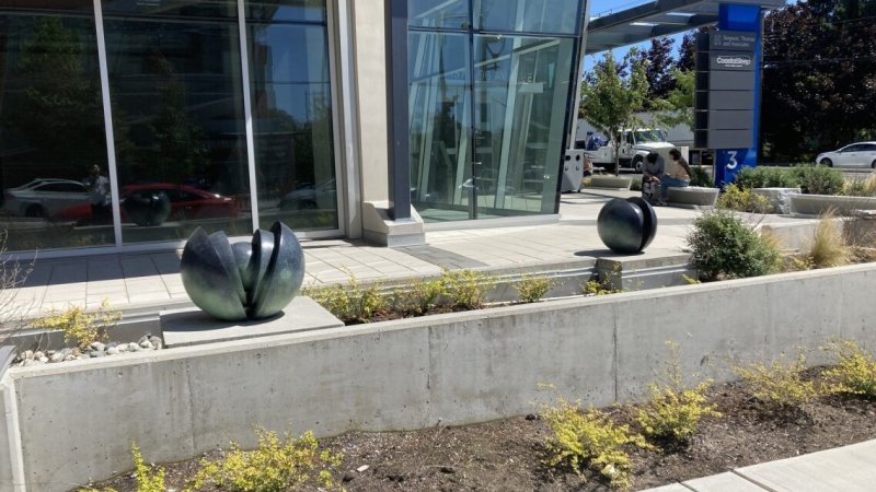 Two abstract sculptures resembling seed pods sit in a modern plaza with shrubs, and a reflective glass building in the background