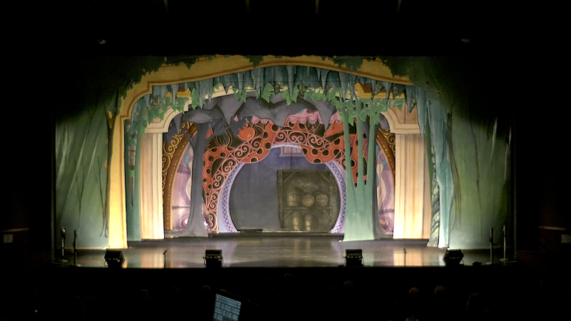 A photograph of a theatre stage that shows numerous backdrop structures, some green, some yellow, and some with flowers falling over the top. Some structures form a rectangular roof, while others are round portals. 