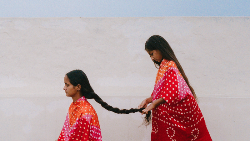 Two children face the left side of the photograph. One child stands behind the seated one to braid her long hair. They both wear bright pink, orange, pink printed clothing, against a white backdrop.