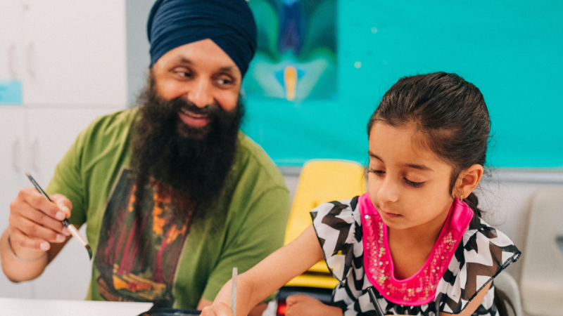 A father looks on with a loving smile at his child, both of them seated at a classroom and holding paintbrushes.