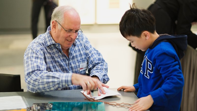 Gallery Event Volunteer Gerald Spie smiles as he shows a stamp activity with a child. They are both intently focusing. 