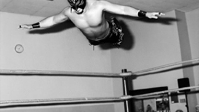 A black and white photograph of a wrestling ring in a room with people seated in chair facing the sides of the wrestling ring. They are looking at an airborne wrestler. The photograph captures the wrestler with arms spread out like wings, legs behind, so they appear to fly. There is a mask covering his skull and face. 