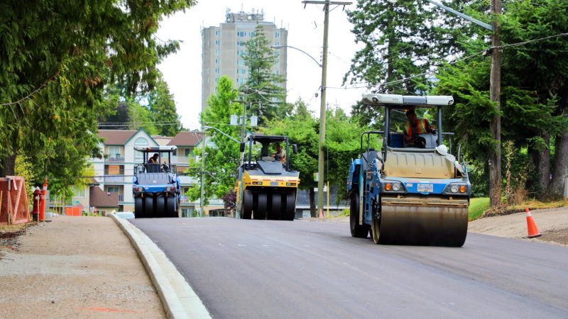  Steamrollers paving a new road with trees and an apartment building in the background.
