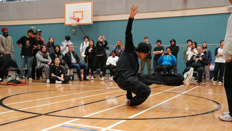 A dancer wearing all black holding a freeze in a gymnasium with an audience around them.