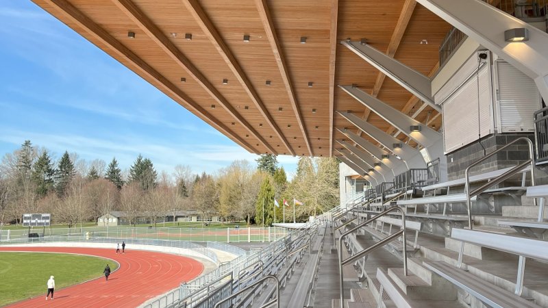 The photo captures an expansive view of a stadium grandstand with a wooden canopy, overlooking an athletic track, under a clear blue sky.