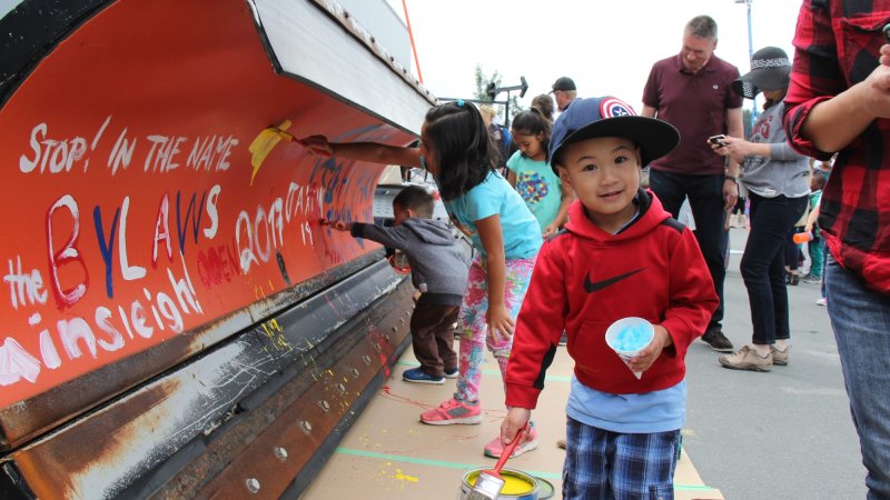 A young boy with a paintbrush in hand happily participating in a community painting event, surrounded by other children and adults.