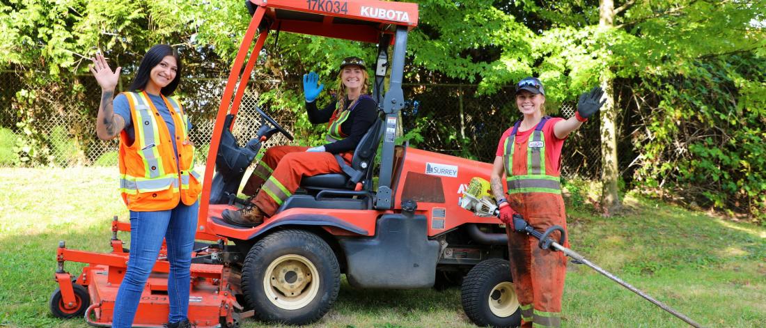 Three female parks staff in front of a tractor are waving