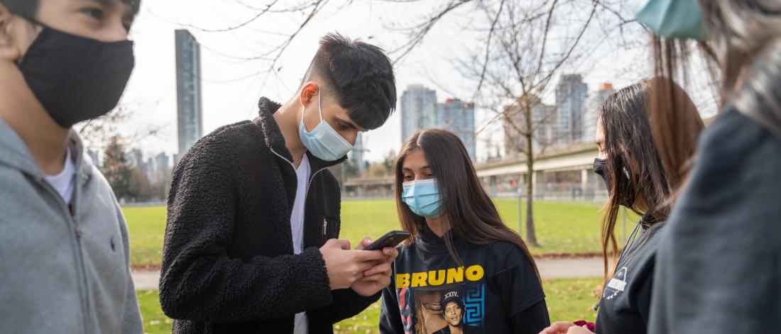 Group of youth wearing face masks, talking and looking at phone.